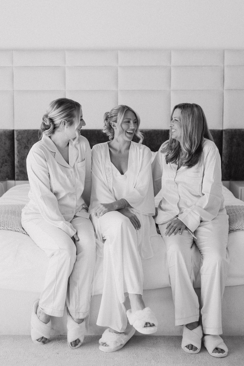 Black and white photo of bride laughing with two bridesmaids in pajamas seated on hotel bed.