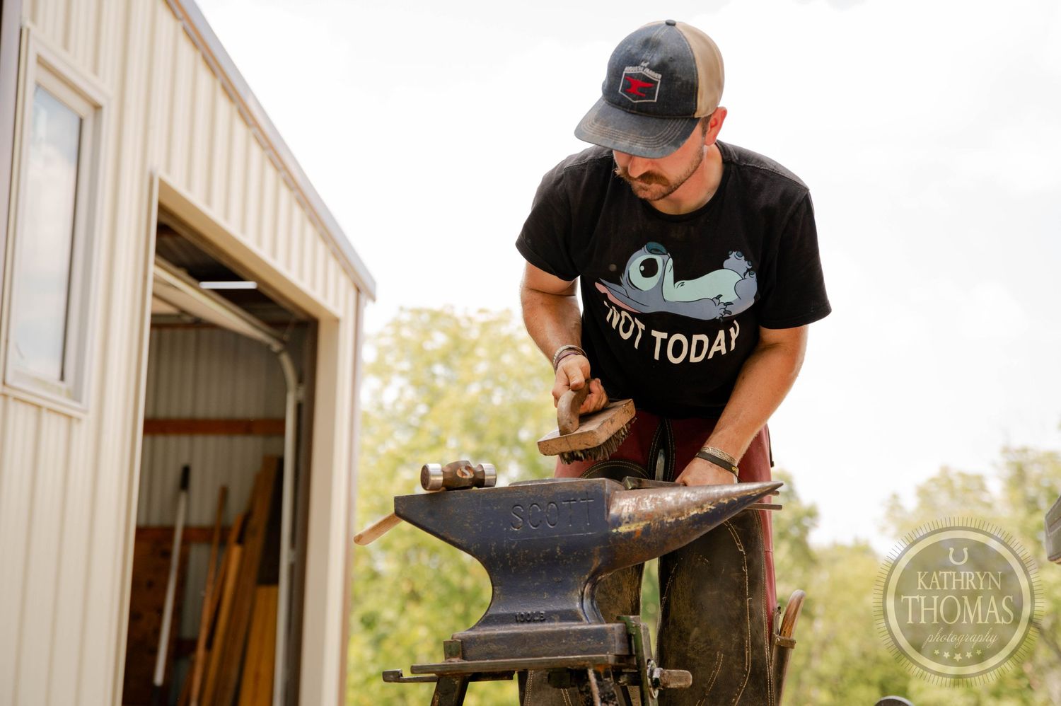 A blacksmith works at an anvil outside a metal workshop wearing protective gear.