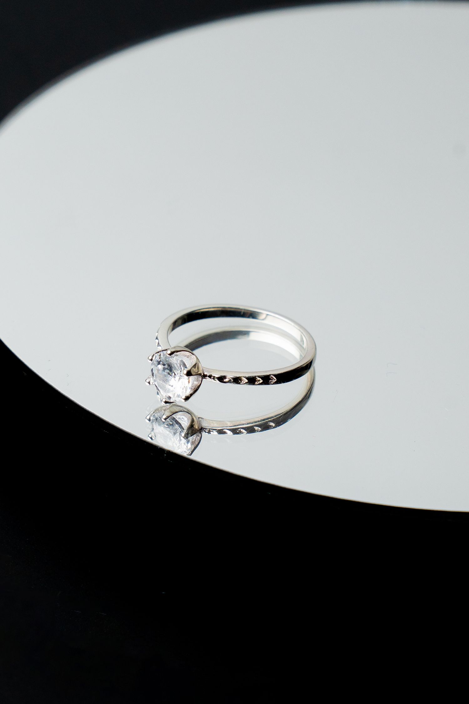 Elegant silver diamond solitaire engagement ring displayed on a reflective white surface against black background.