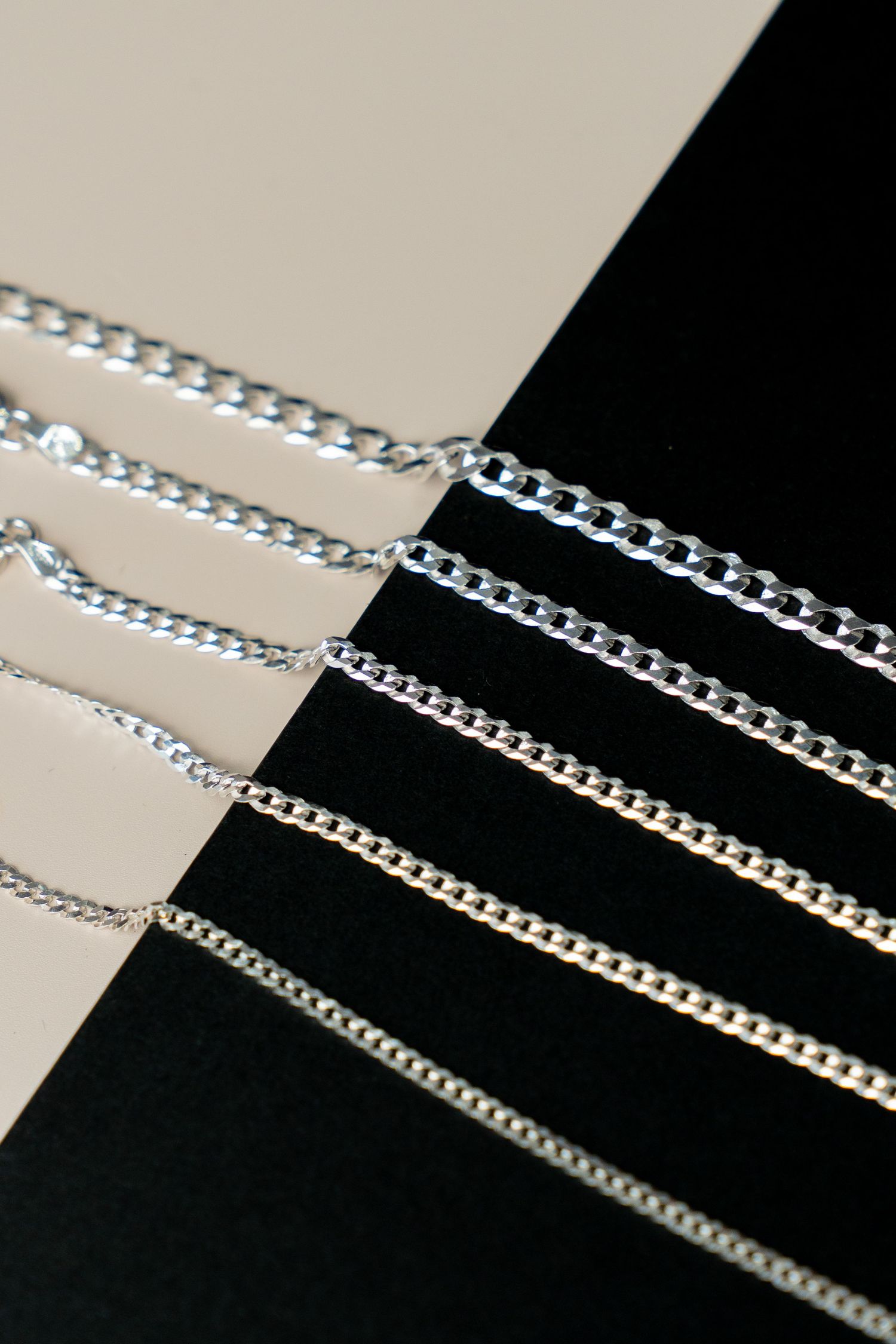 Multiple silver curb chain necklaces displayed diagonally on a black and beige background.