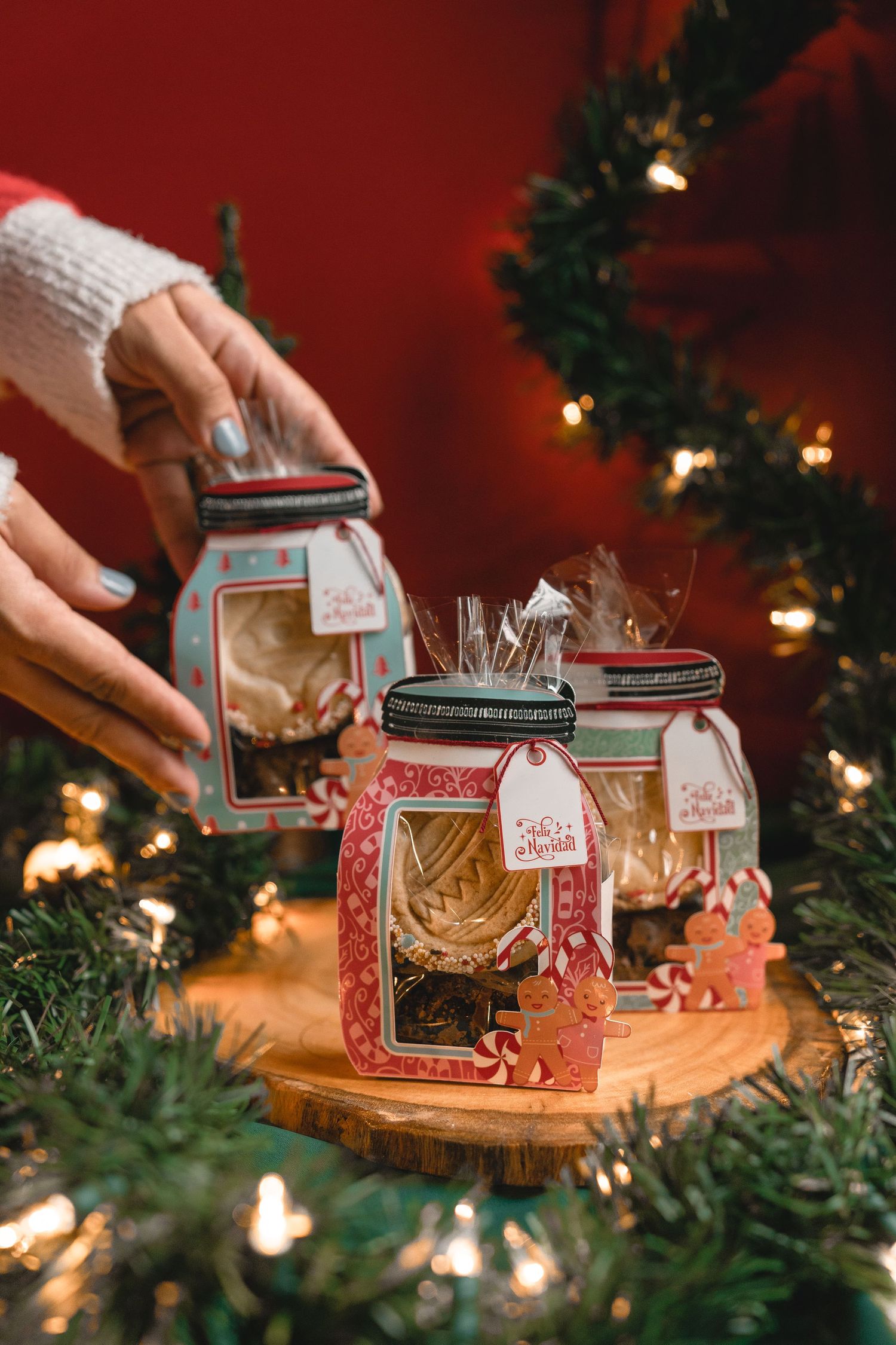 Decorative glass jars with lights and greenery arranged on wooden surface with Christmas tree in background.