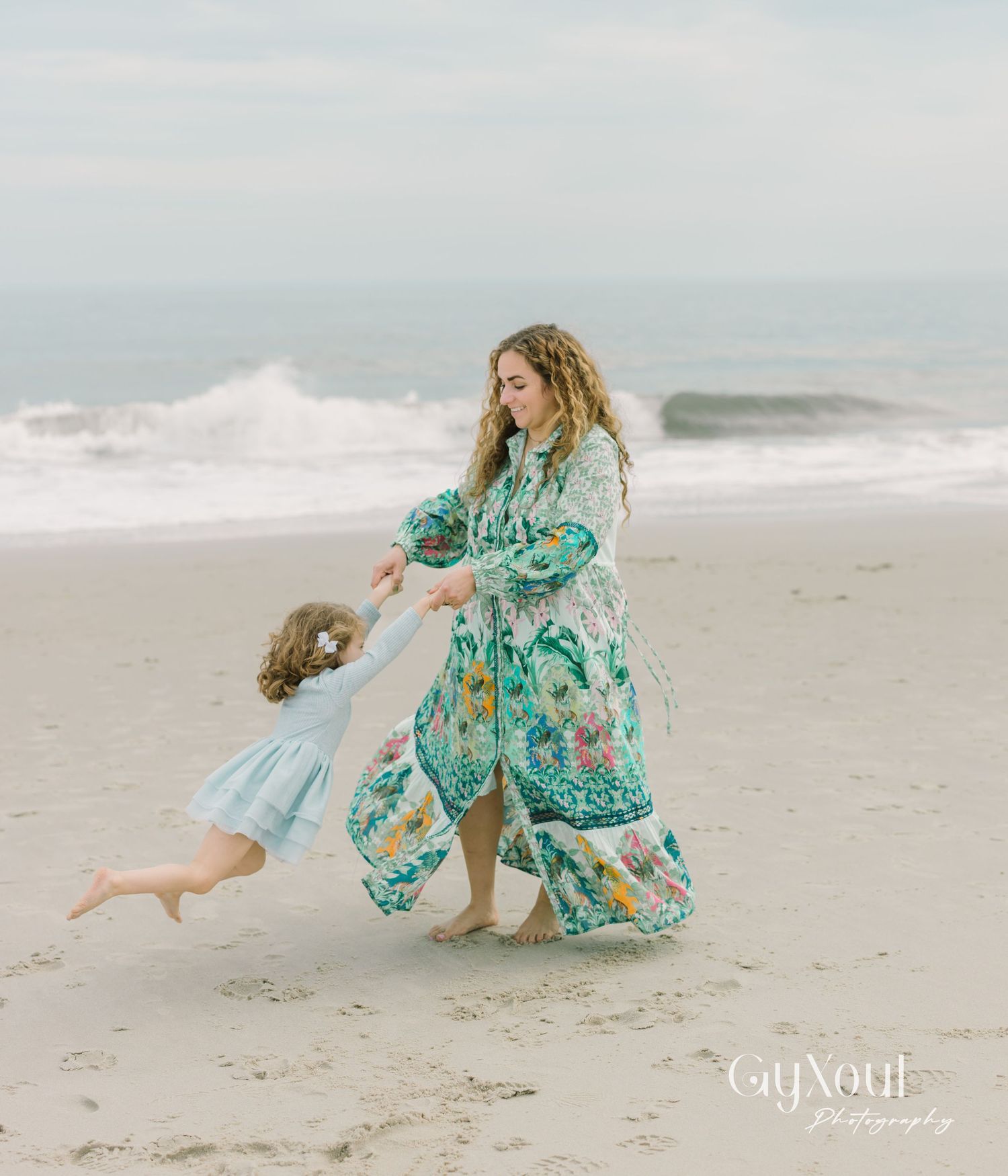 A parent and child in matching floral teal dresses dance together on a misty beach with crashing waves in the background.