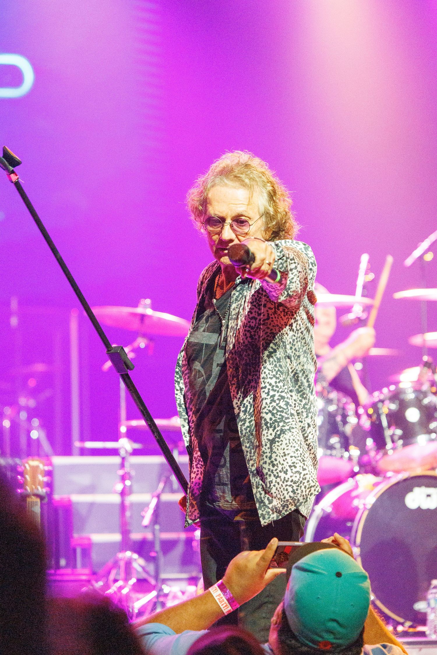 Performer in sparkly silver jacket sings on stage with purple lighting and drum kit visible in background.