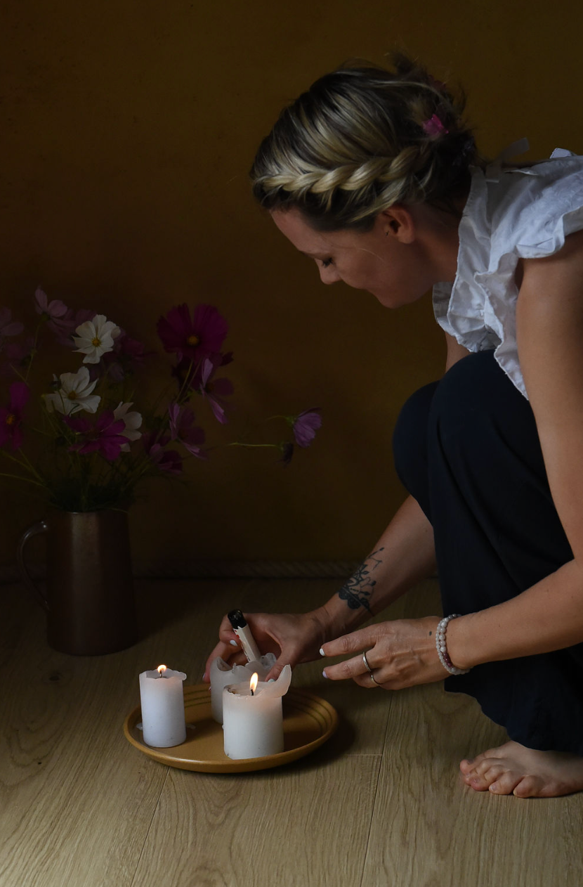A person in a white blouse lights candles on a wooden floor, with purple flowers in a vase creating a peaceful atmosphere.