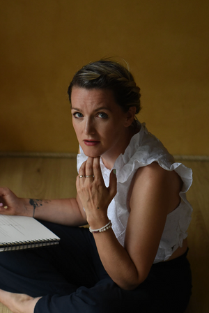 A person in a white ruffled blouse and dark pants sits thoughtfully at a desk with a notebook.
