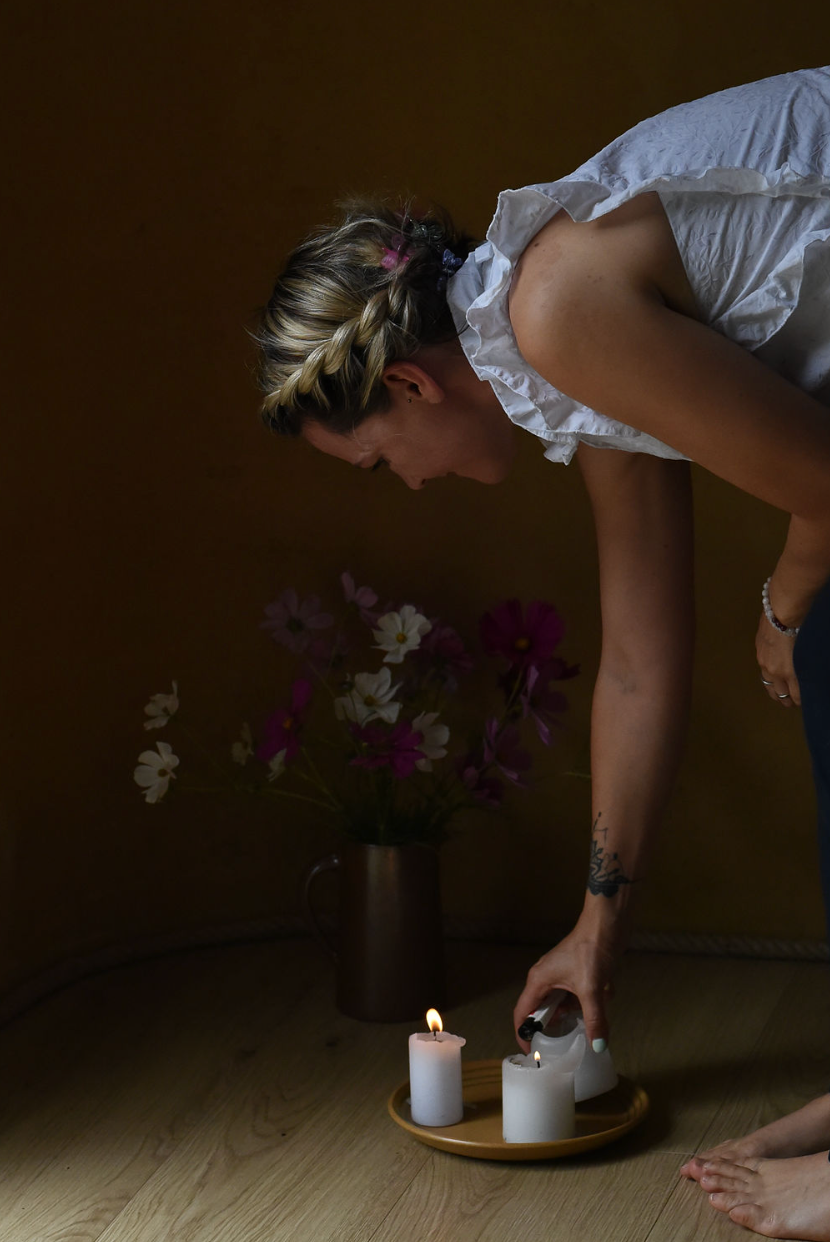 Person in white top bends down to arrange candles on wooden floor beside floral arrangement in dark room.