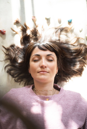 Person wearing purple sweater lies down with dark hair splayed out dramatically against white background.