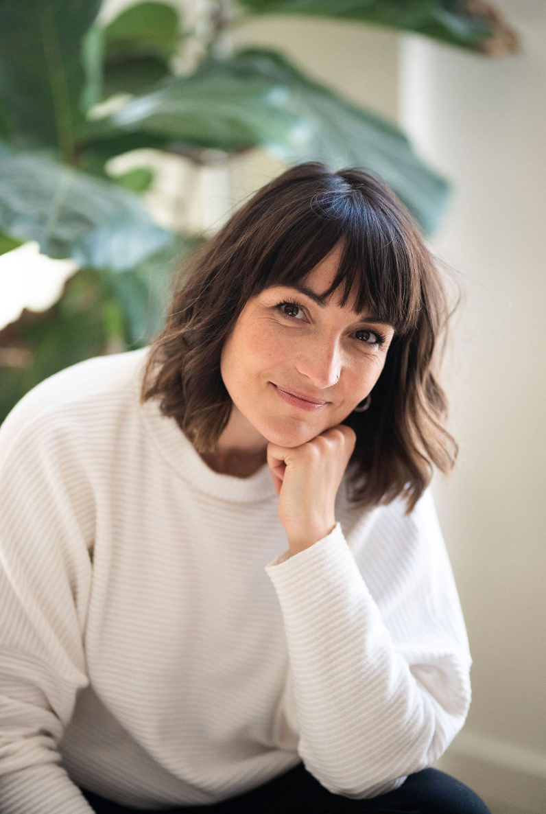 A person with dark bangs and shoulder-length hair rests their chin on hand while wearing a cozy white sweater near indoor plants.