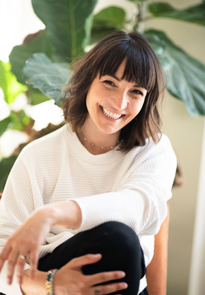 Someone wearing a white knit sweater smiles warmly while sitting casually near a green houseplant in natural lighting.