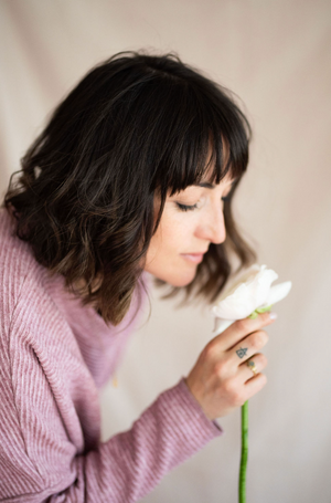 Side profile view of person in pink sweater gently smelling a white flower against light background.