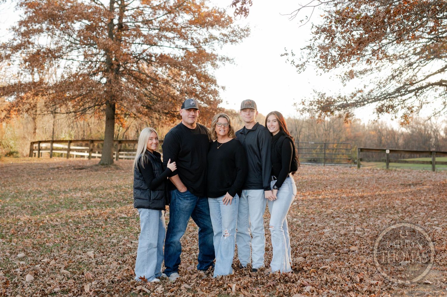 Family of five stands close together on fallen leaves in an autumn park setting.