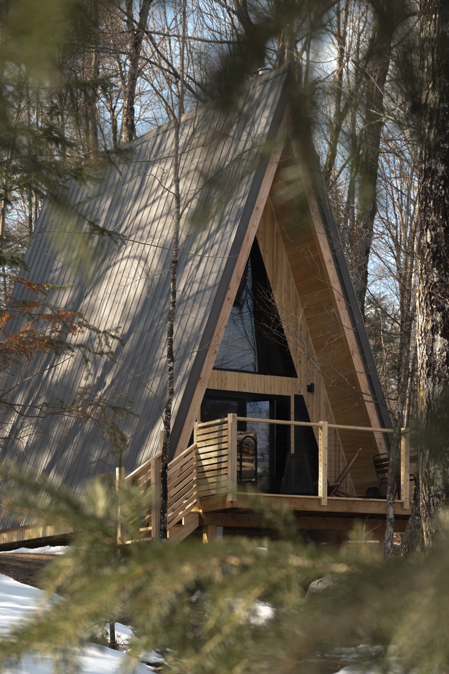 A rustic A-frame cabin with wooden exterior and deck nestled among bare winter trees in a snowy forest.