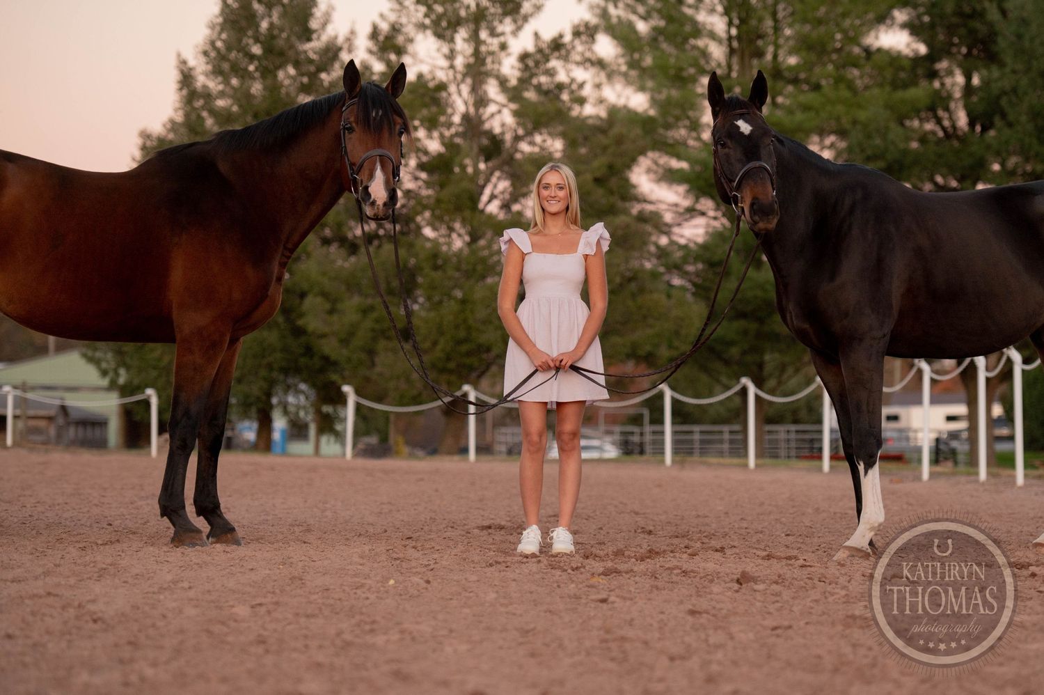 Person in white dress standing between two majestic horses in a fenced arena at sunset.