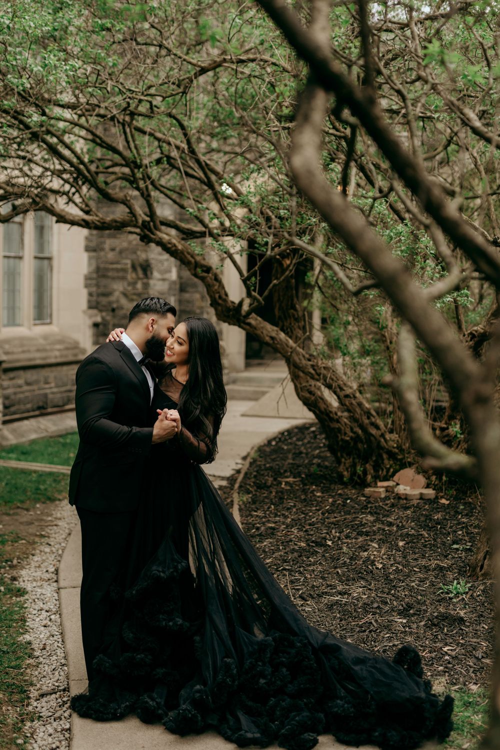 A romantic couple in black attire embrace under twisted tree branches near a stone building.