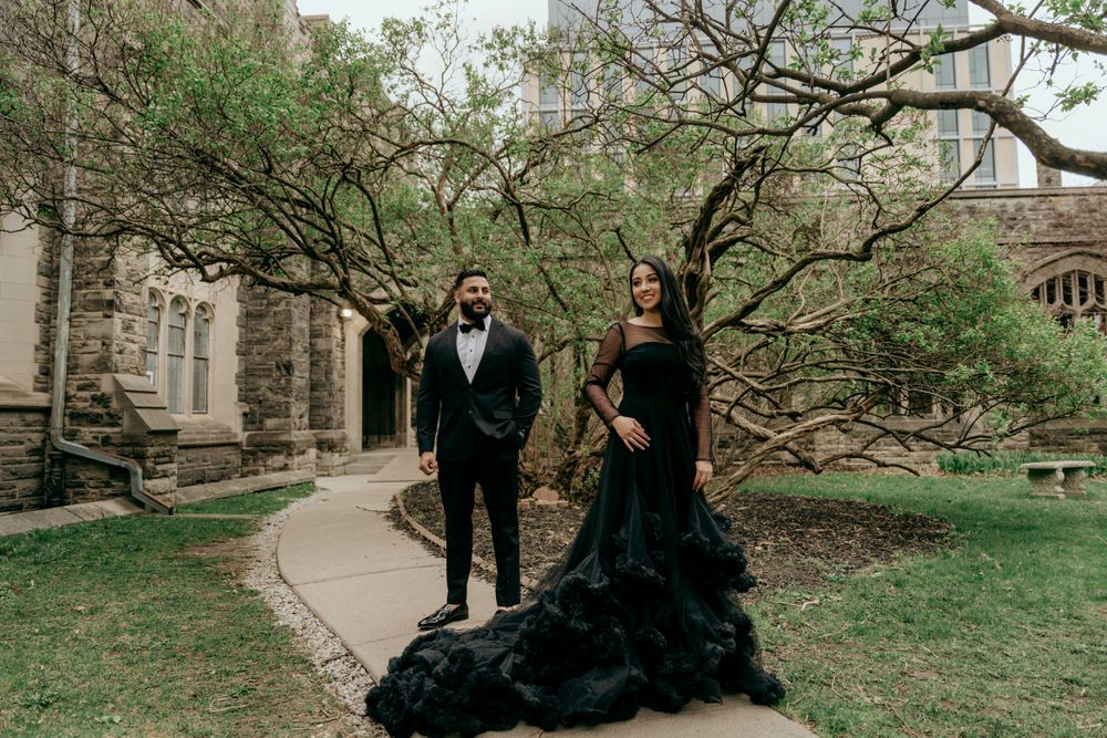 A couple in formal black clothing poses on a garden path near gothic architecture.