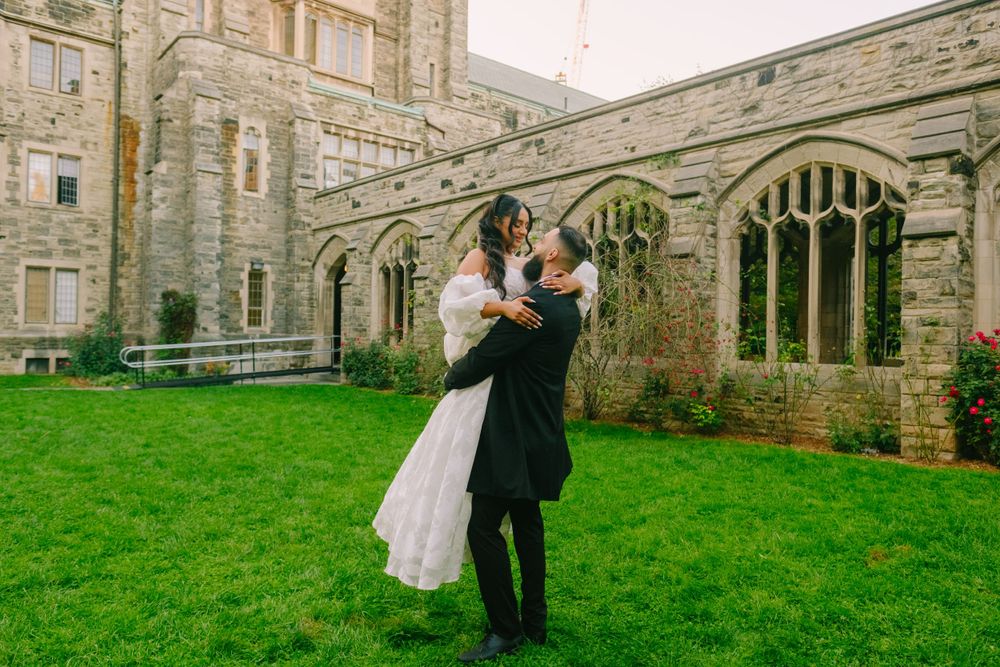 A couple embraces in formal wedding attire on a lush green lawn beside a historic stone church with Gothic architecture.