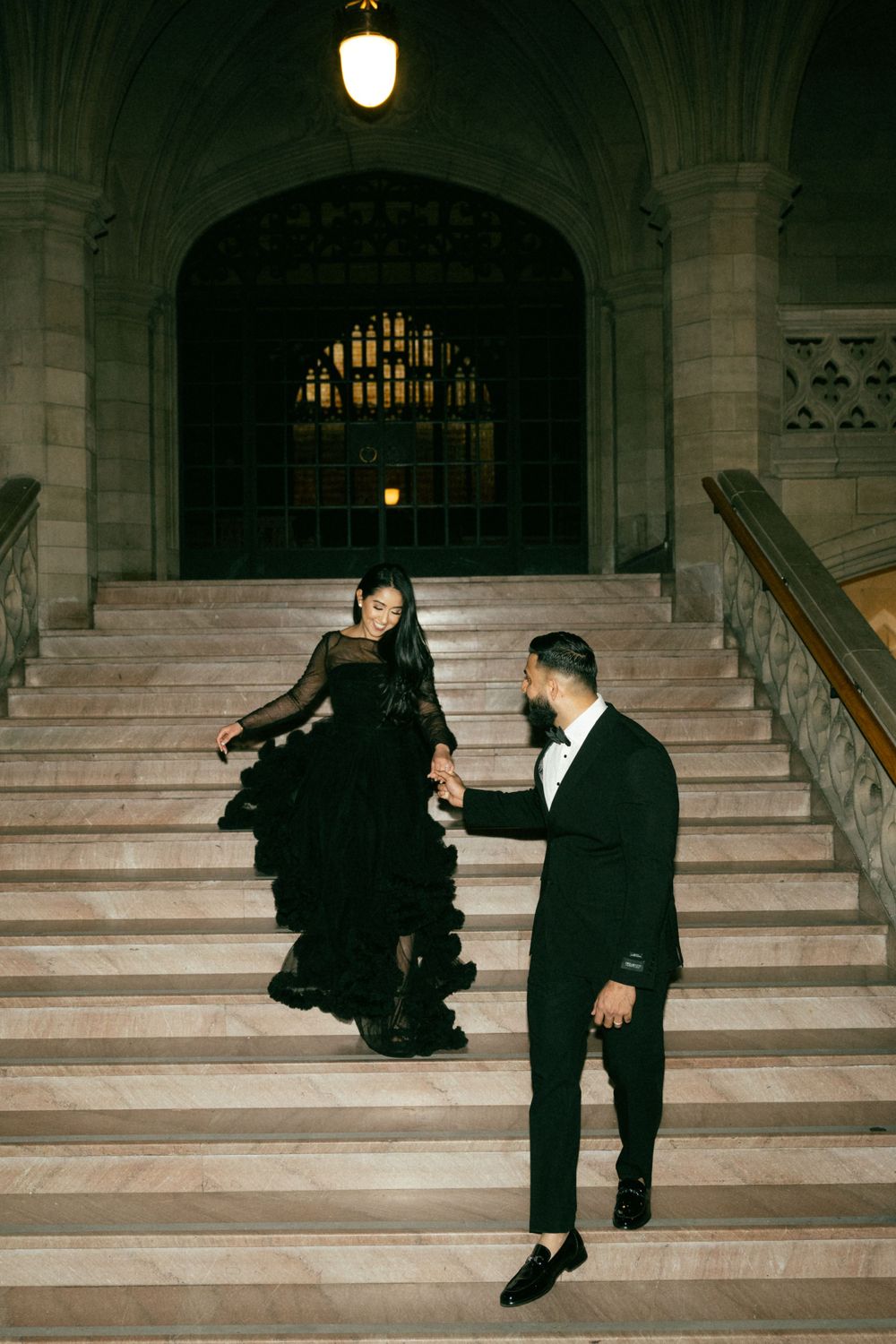 Elegant couple in formal attire descending grand stone staircase in vintage architectural setting with arched ceiling.