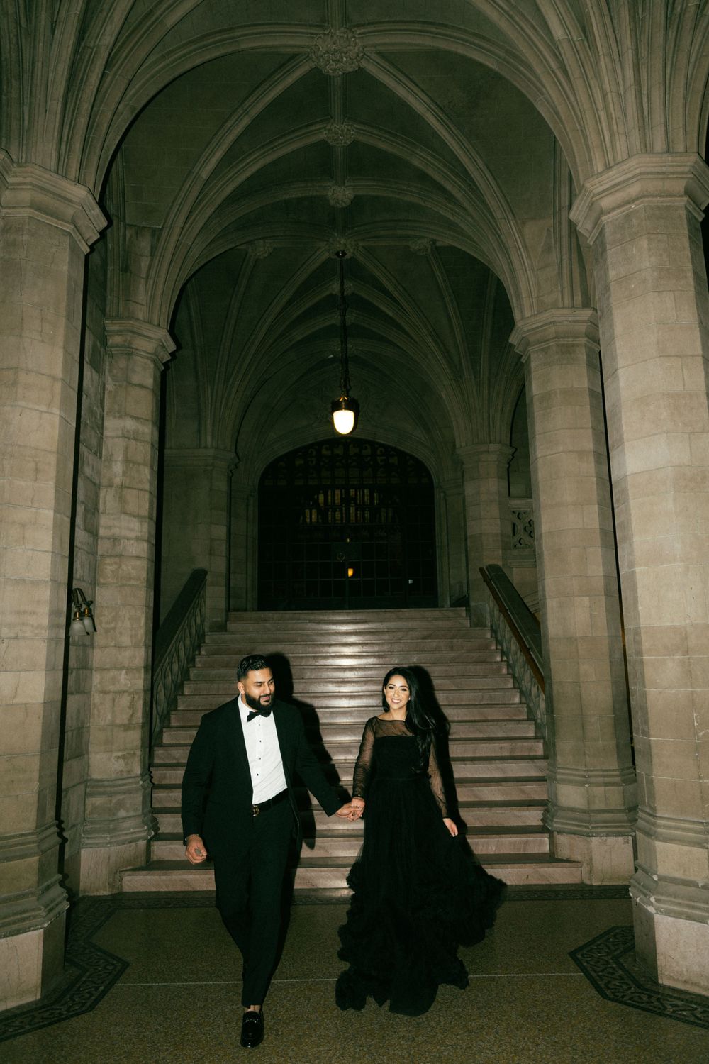 Elegant couple in formal black attire descending historic stone staircase in Gothic arched hallway at night.