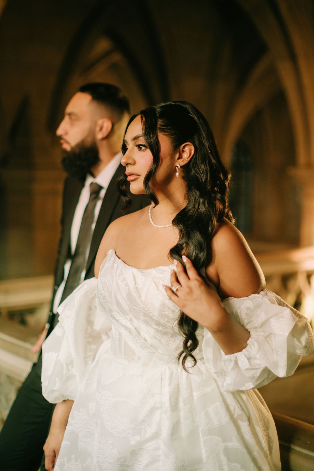 Elegant couple in formal wedding attire pose in front of gothic arched windows with warm lighting.