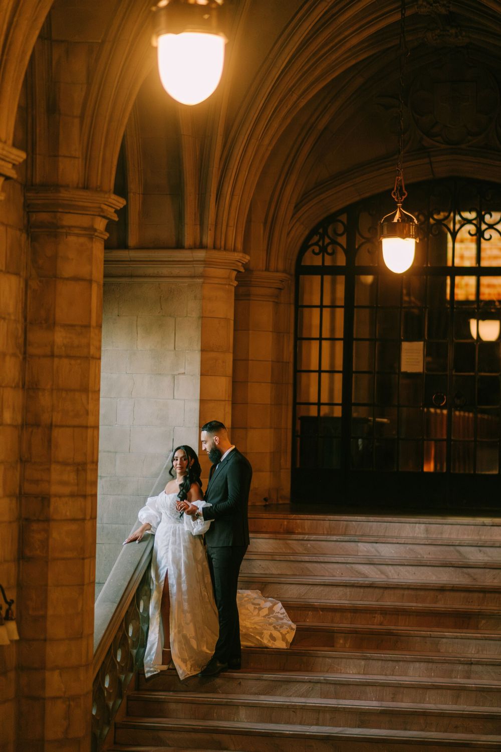 A romantic wedding couple stands on stone steps at dusk, illuminated by warm vintage pendant lights in an elegant archway.