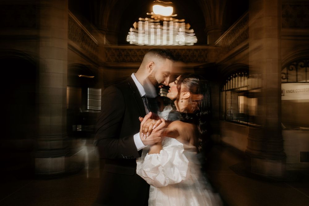 A romantic wedding couple shares an intimate dance moment beneath an elegant crystal chandelier in a grand ballroom.