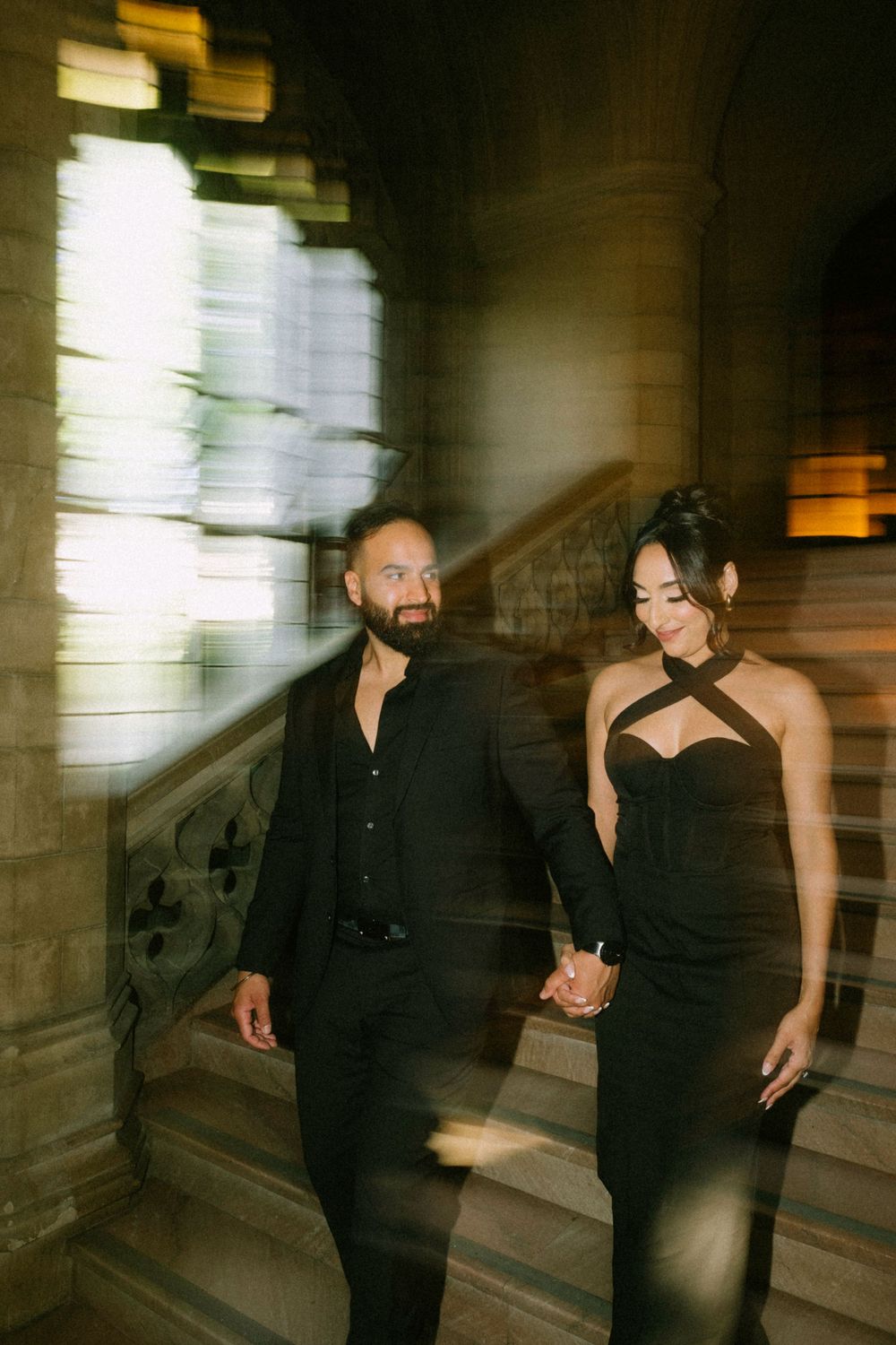 A couple in elegant black formal wear descends a grand stone staircase in a dramatic, dimly lit interior.