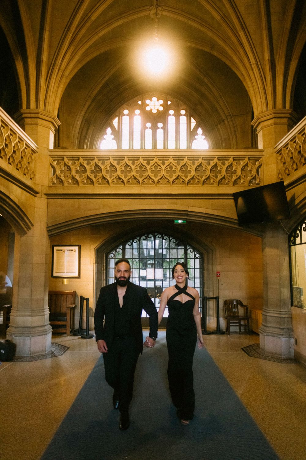 Two elegantly dressed people walk down a grand gothic hallway with vaulted ceilings and decorative stonework.