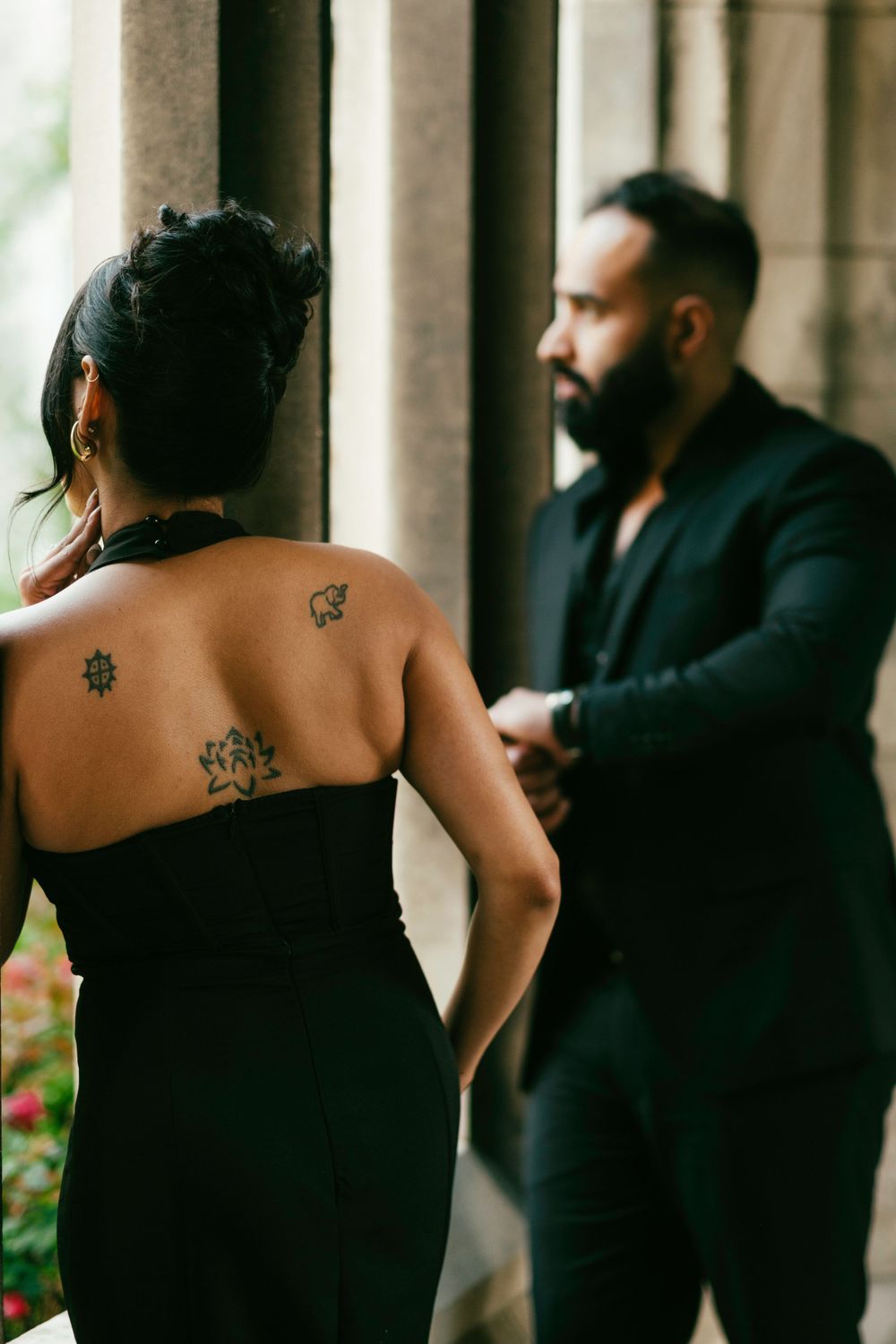A couple in formal attire stands in a colonnade, with the woman wearing a black dress showcasing tattoos on her bare back.
