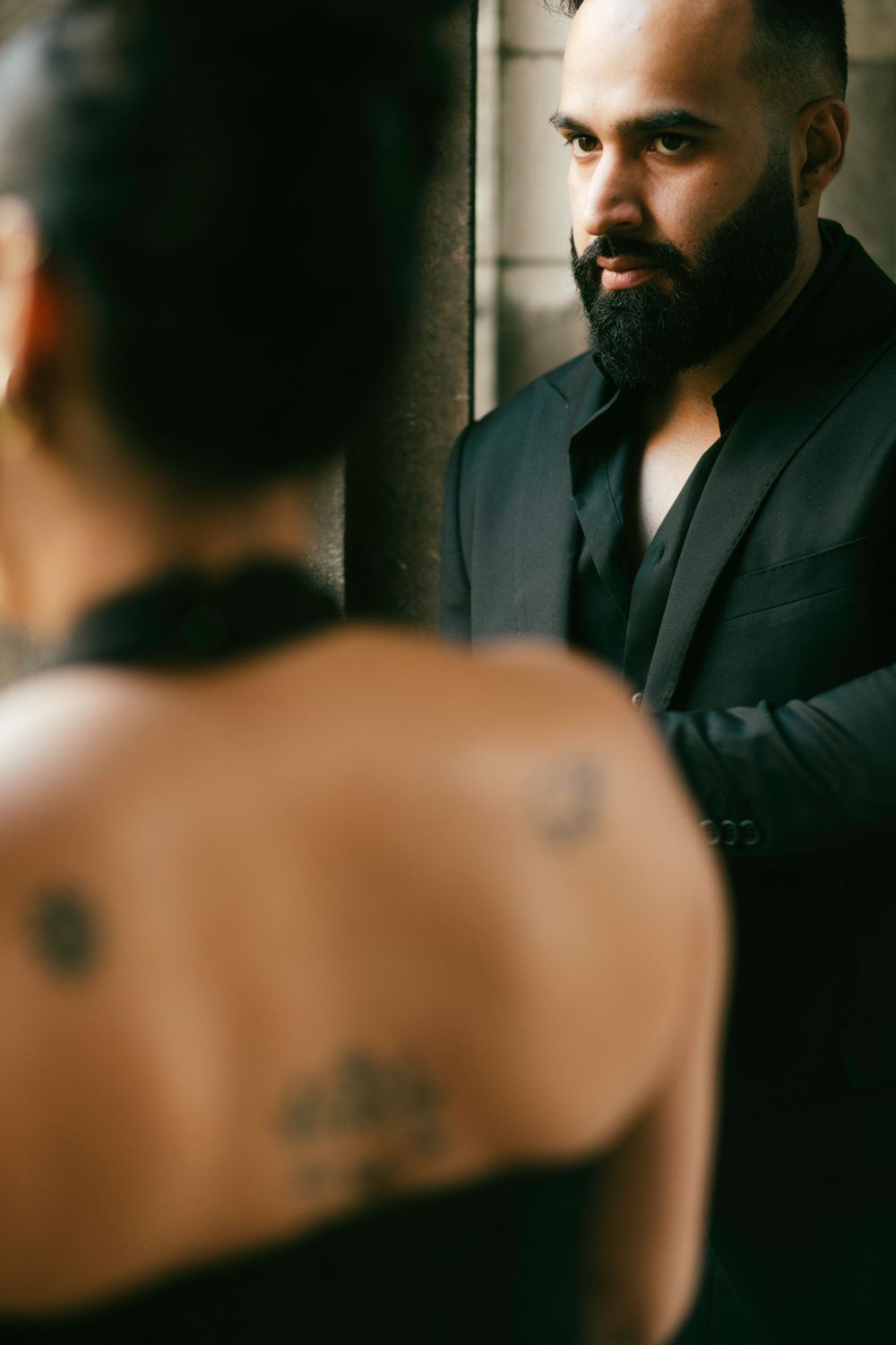 Person with dark beard wearing black suit jacket gazes thoughtfully into mirror reflection in atmospheric lighting.