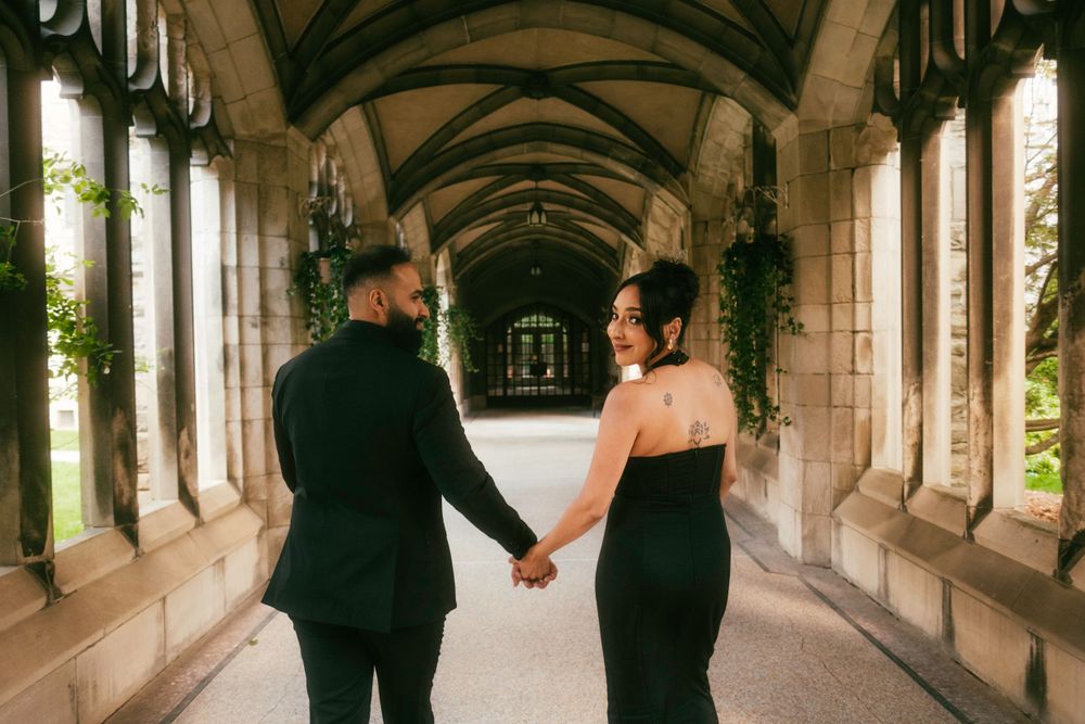 A couple in formal black attire holds hands while walking through an elegant stone archway corridor.