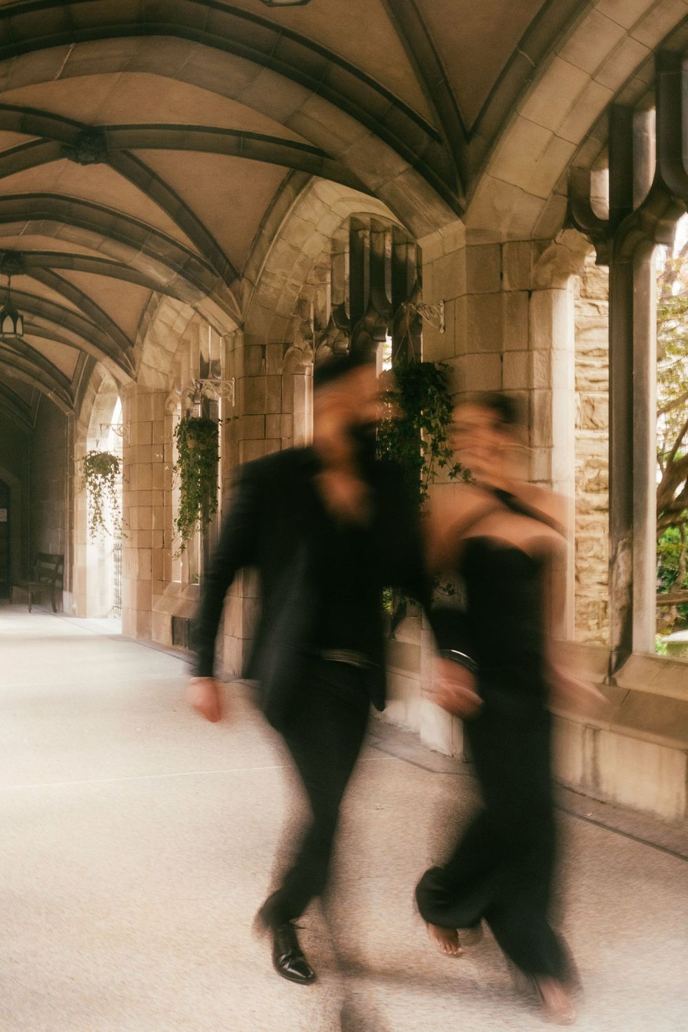 Blurred motion silhouettes walking through a gothic stone archway corridor with vaulted ceilings and natural light.