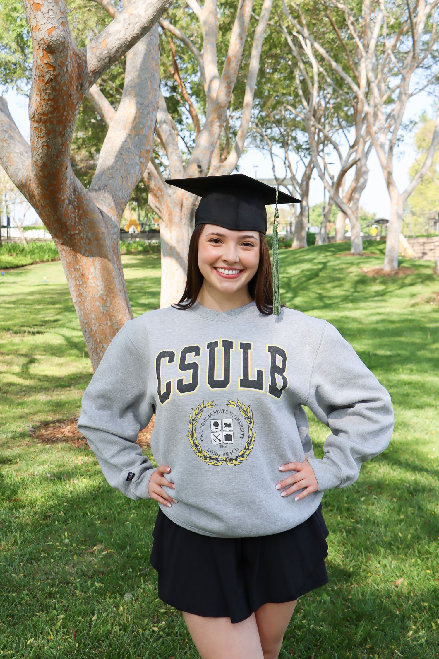Graduate wearing CSULB sweatshirt and cap posing in campus garden.