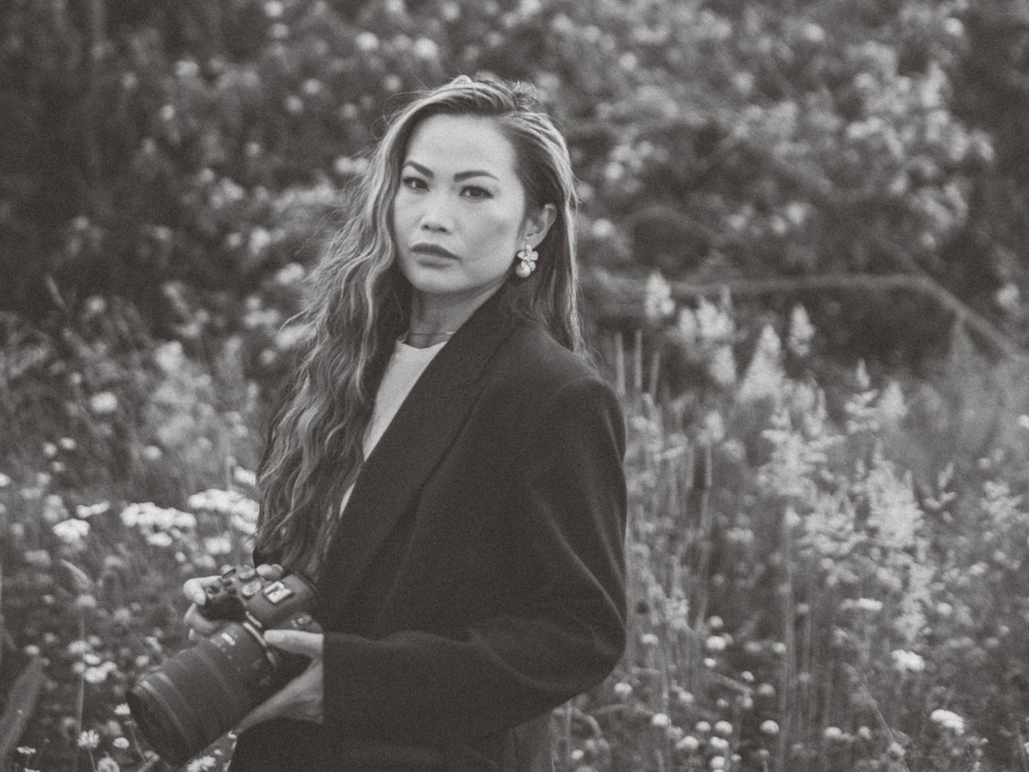 Black and white portrait of person with long wavy hair wearing dark blazer in garden setting.