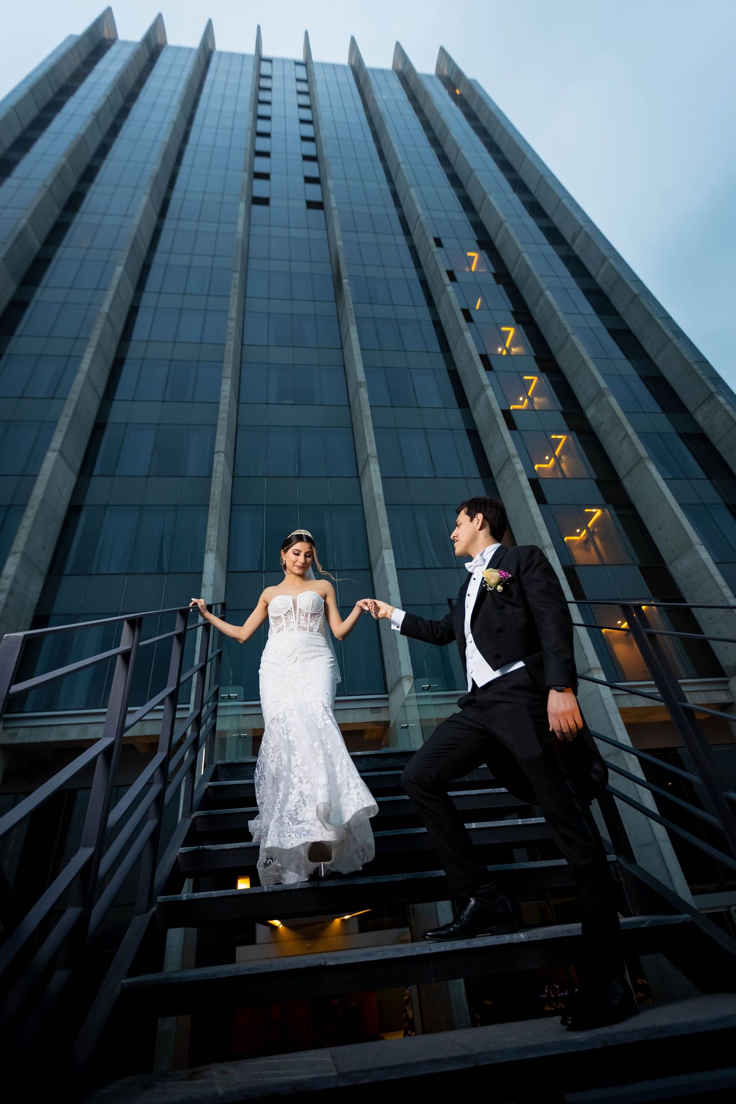 Dramatic wedding photo of newlyweds posing on modern glass building staircase at dusk.