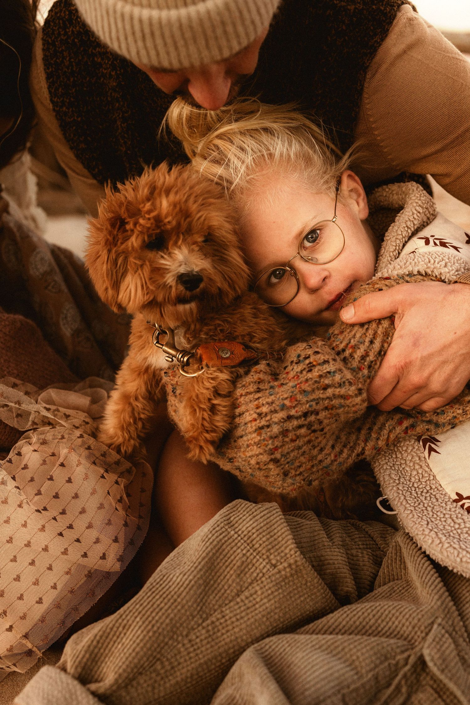 A small child and a brown teddy bear-like dog cuddle together in soft blankets with warm lighting. Javea family photographer