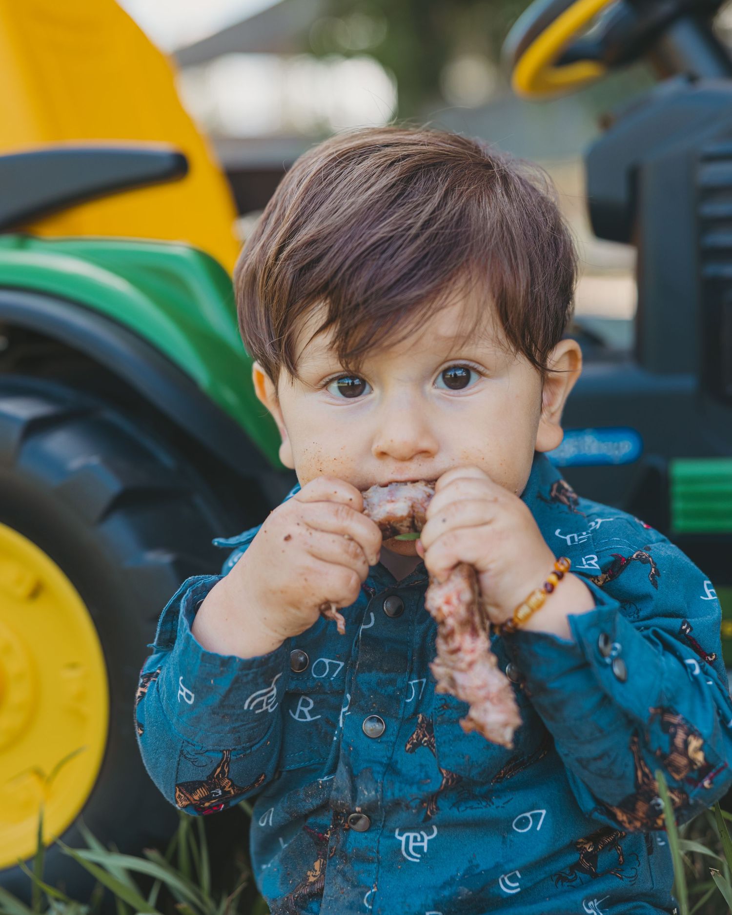 Cute toddler boy in blue shirt eats food in front of a green and yellow toy tractor outdoors.