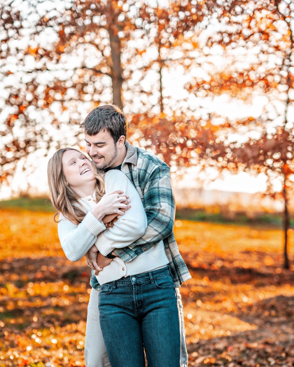 Romantic couple embraces in autumn setting with orange fall foliage and golden sunlight in background.
