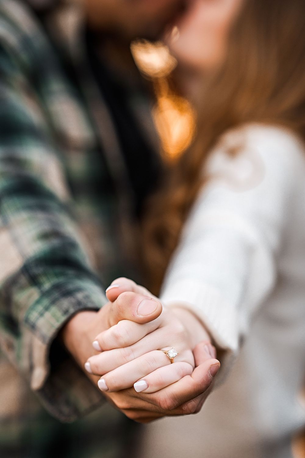 Close-up photo of couple holding hands with engagement ring visible, wearing plaid shirt and white sweater.