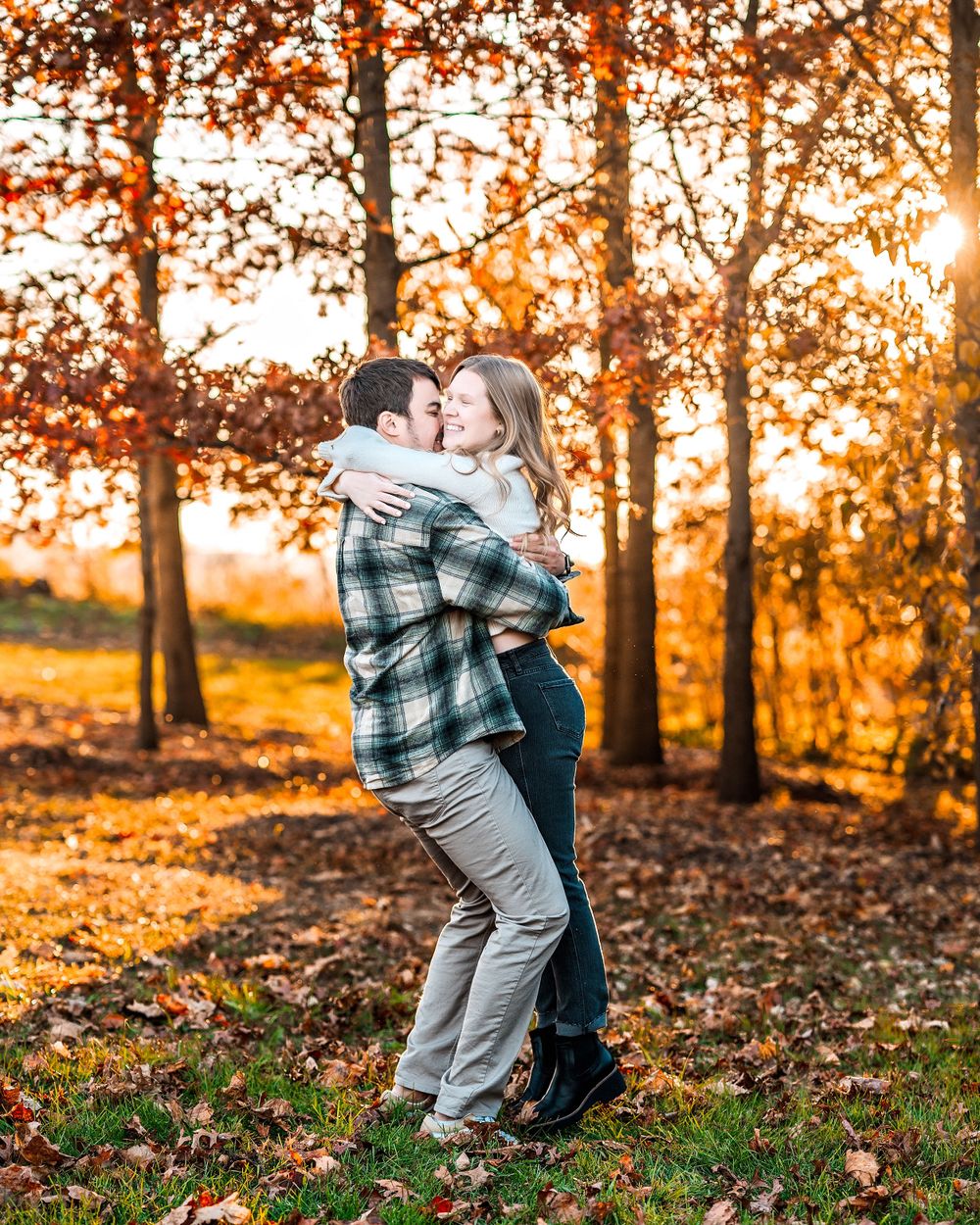 A couple embraces in front of autumn trees during golden sunset, with fallen leaves scattered on the ground.