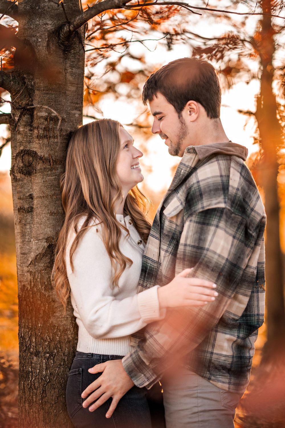 A couple embraces among autumn trees during golden hour sunset as warm light filters through branches.