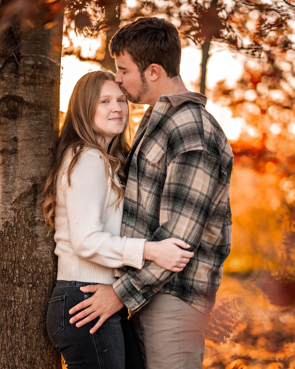 Intimate couple shares tender moment against tree trunk with warm autumn sunset glowing behind them.