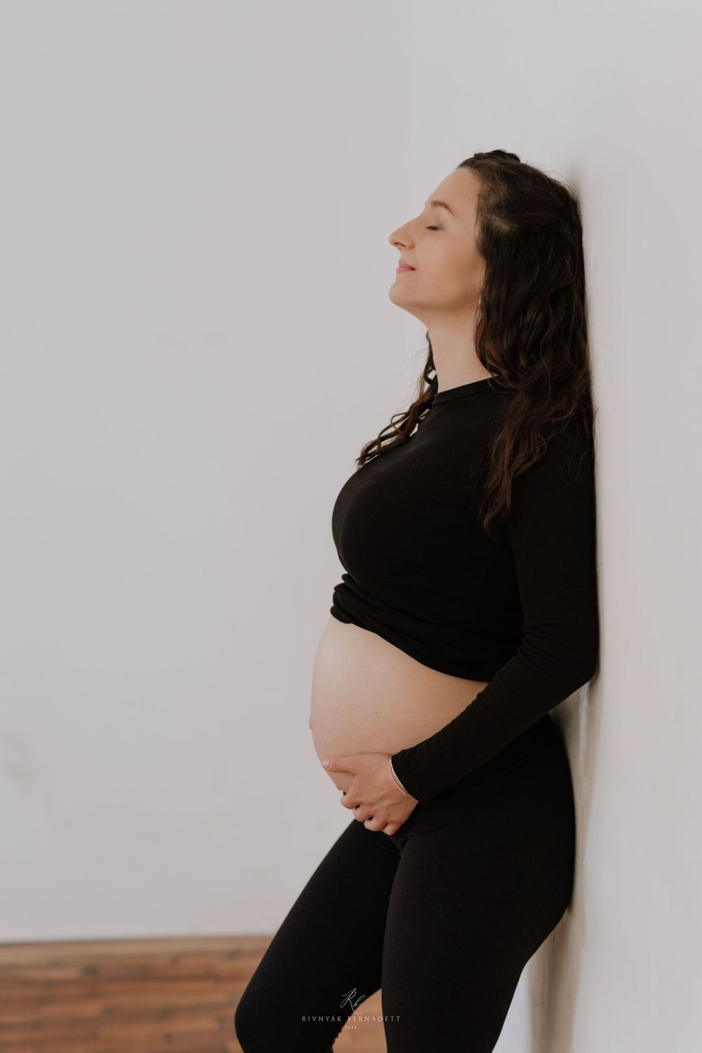 Expectant mother in black attire poses against white wall in serene maternity photo.