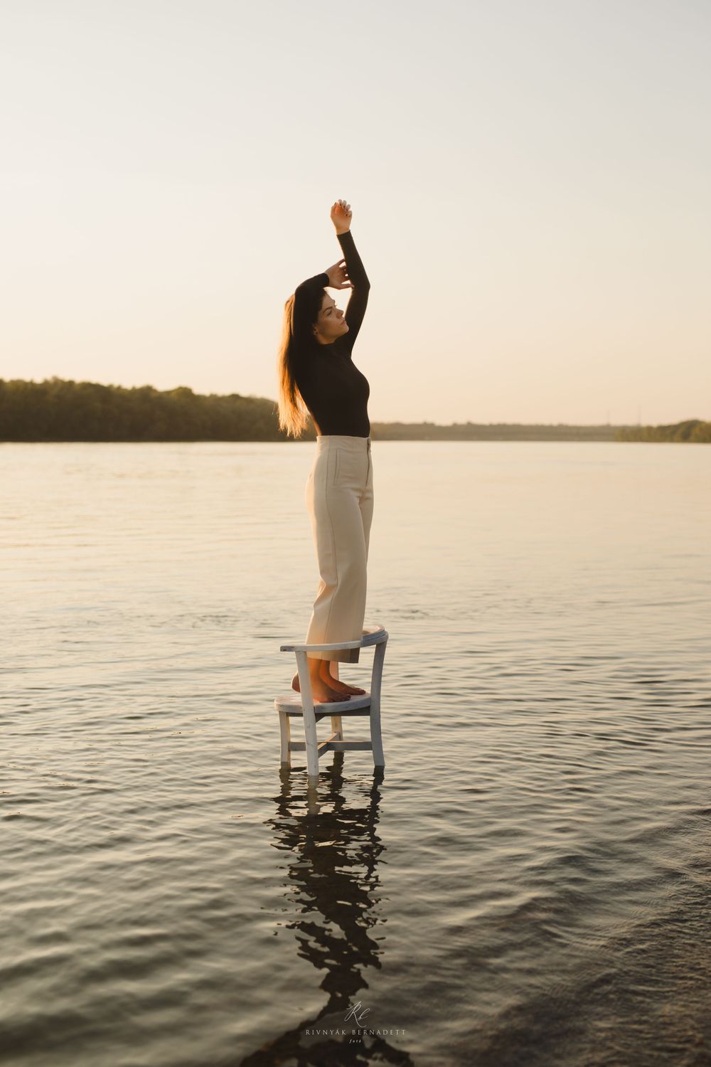 Silhouetted figure standing on chair in water during sunset.