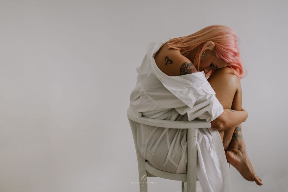 Person draped in white fabric sitting curled up on a chair in a minimalist setting.