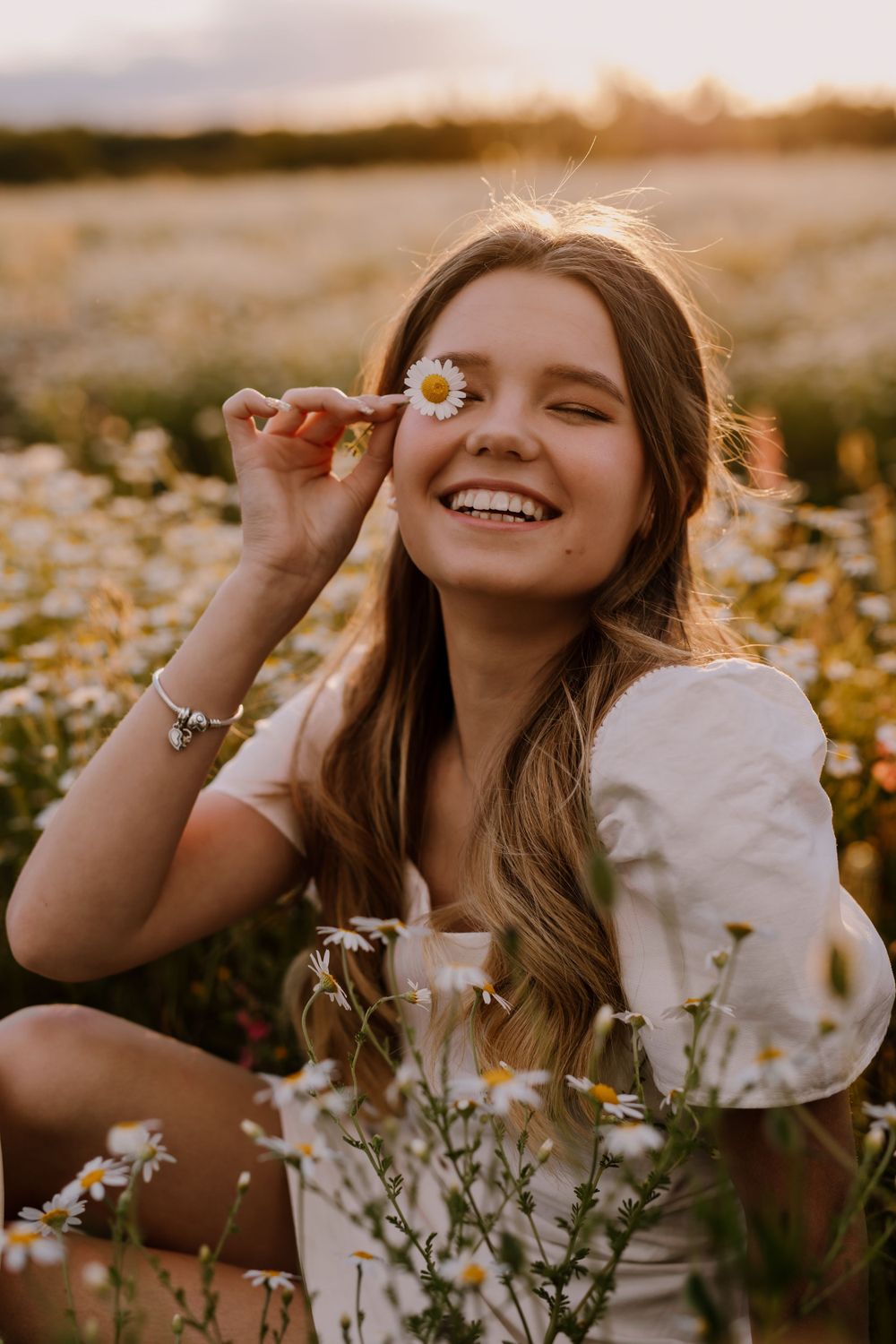 Person in white dress sitting among blooming daisies at golden hour.