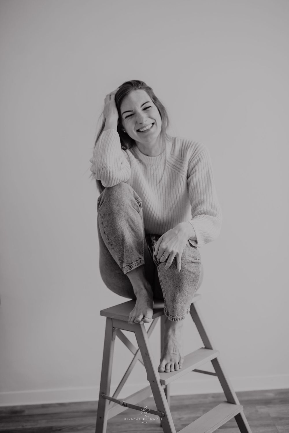 Casual portrait of someone sitting on a wooden stool against a grey background.