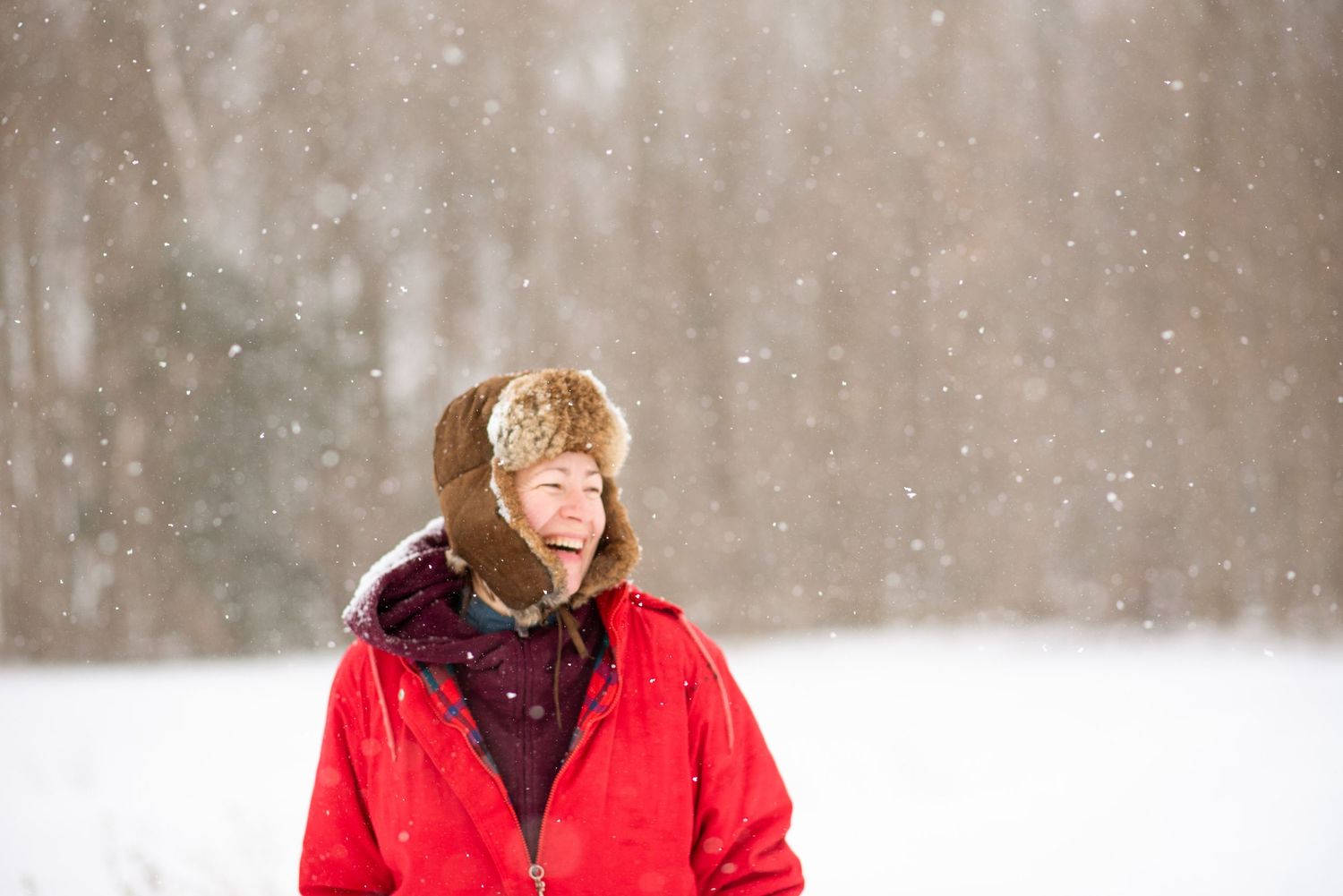 Person in a bright red winter coat and fur hat smiles while standing in falling snow against a white background.