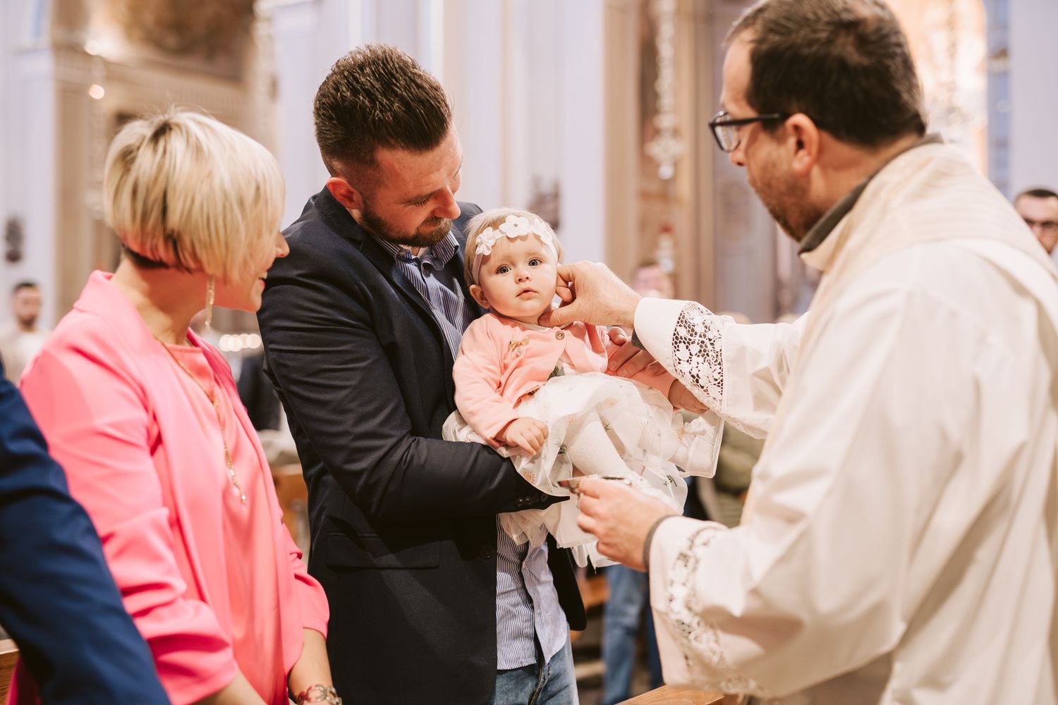 Bambina in chiesa riceve uno dei riti del Battesimo dal prete mentre il papà la tiene in braccio e la mamma le sorride accanto.