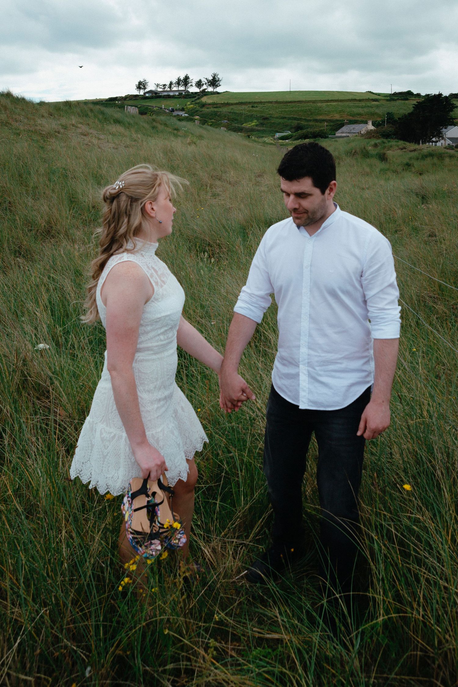 A couple stands together in a grassy meadow, with the woman wearing a white dress and the man in a white button-down shirt.