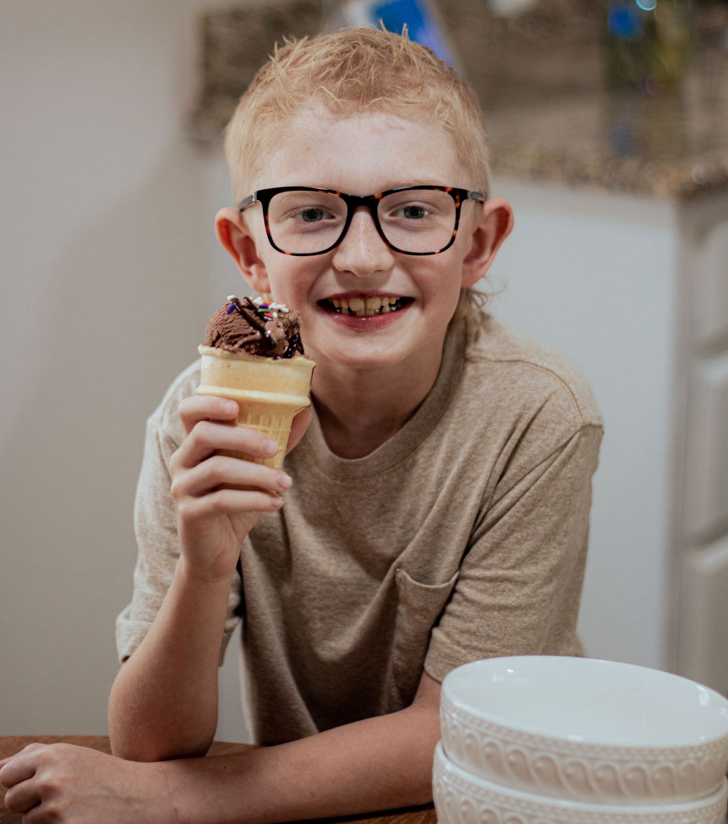 A young student in glasses and a tan sweater smiles while holding an ice cream cone.